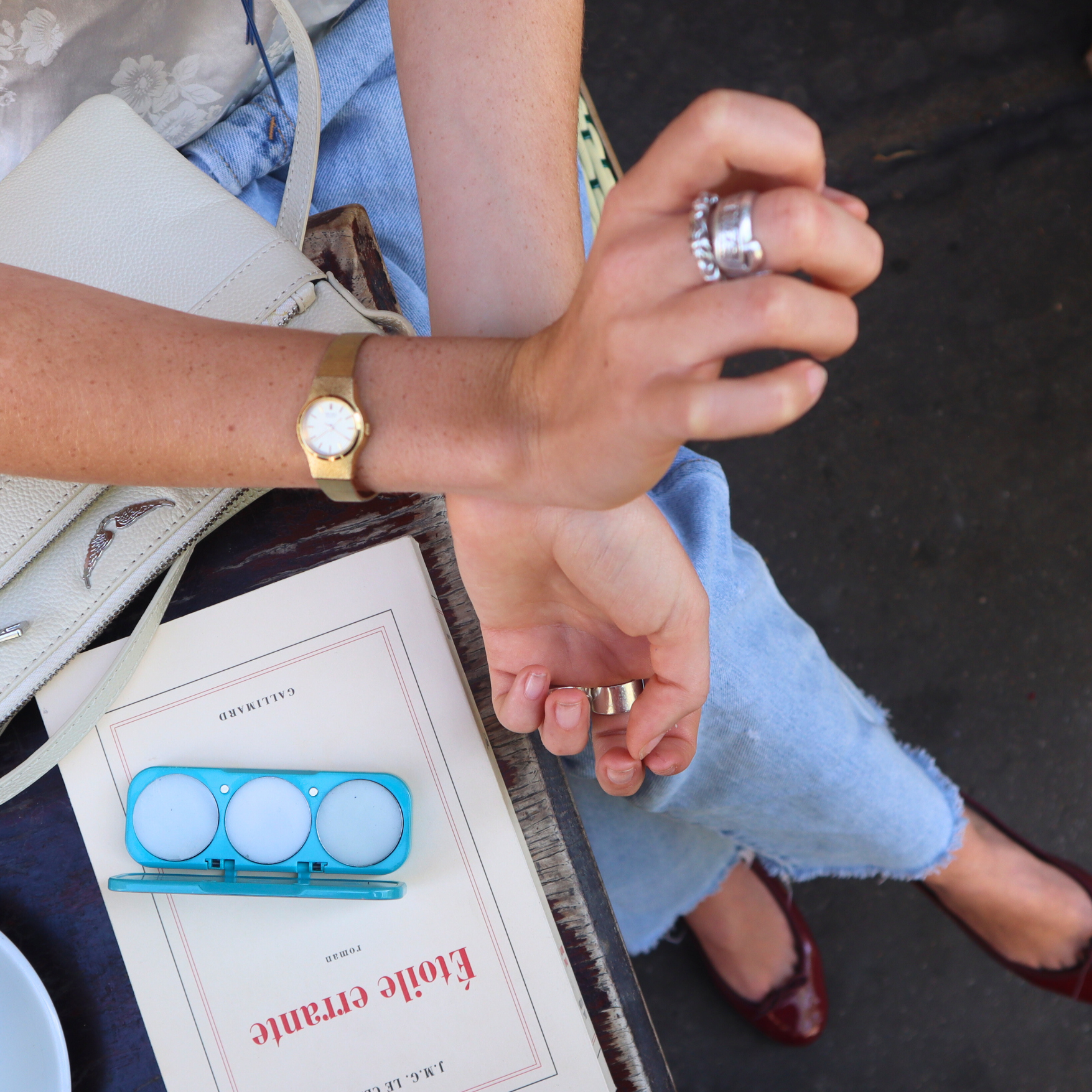 Person holding a book with a blue object on top, wearing a gold watch and ring, applying ema fragrance solid perfume to her wrist.