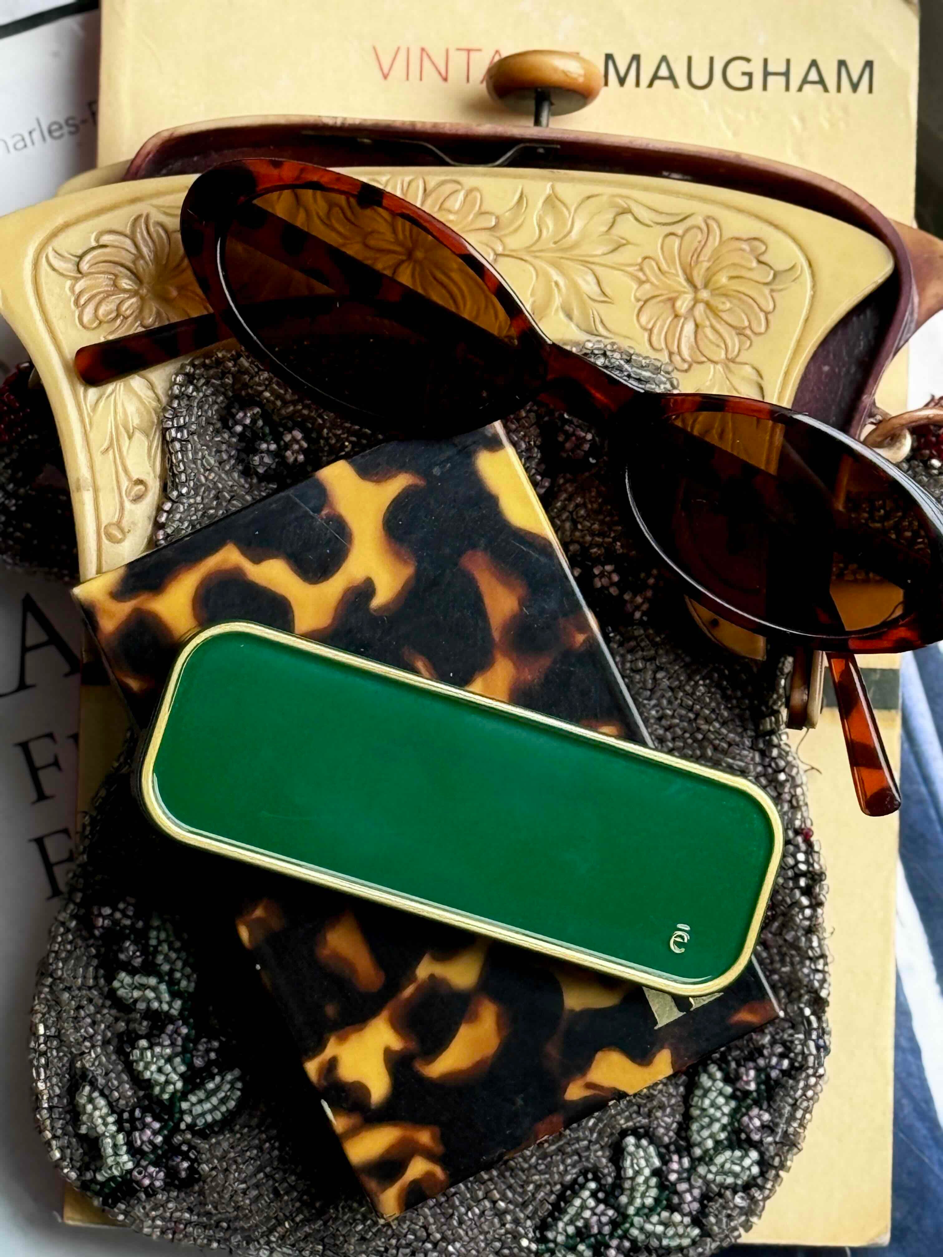 Tortoiseshell sunglasses and green ema fragrance solid perfume compact on a textured surface with 'Vintage Maugham' branding.