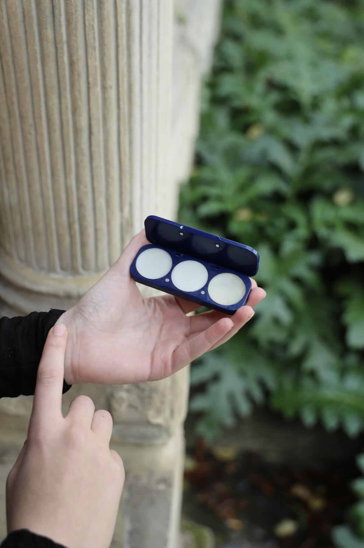 Hand holding solid perfume compact and applying perfume to wrist, against a blurred outdoor background