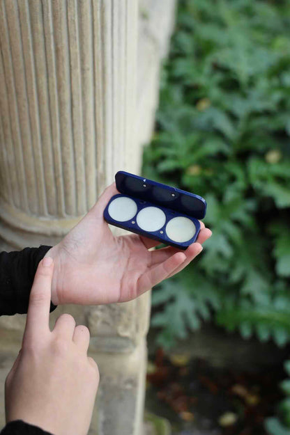 Hand holding solid perfume compact and applying perfume to wrist, against a blurred outdoor background
